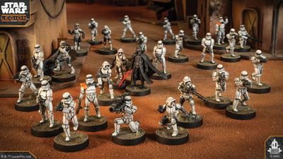 A group of Star Wars miniatures features several Stormtroopers in white armor, positioned in a desert-like environment with a backdrop of a weathered building. Among them stands a prominent figure in black armor, holding a red lightsaber, likely representing Darth Vader. The scene is detailed with various poses of the troopers, some aiming weapons, while others appear to be in action, all set on circular bases with textured surfaces.