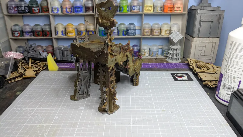 An Orc Lookout model is prominently displayed on a crafting mat, showcasing intricate details and a weathered appearance. In the background, a shelf holds various paint bottles, while additional terrain pieces and tools are visible on the workspace. The scene captures a creative environment focused on tabletop gaming and model building.
