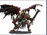 A large, intricately detailed statue of Angron, the Daemon Primarch of Khorne, stands prominently. He features massive, bat-like wings and a fierce expression, with a head that appears relatively small compared to his muscular body. Adorned with skulls and ornate armor, he wields a large sword in one hand and a weapon in the other, standing on a textured base that enhances the dramatic effect of the figure. The color palette includes deep reds, blacks, and metallic tones, emphasizing his menacing presence.