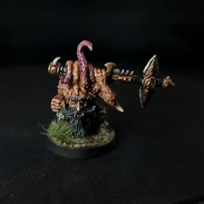 A miniature figure of a Chaos Dwarf with a muscular build, holding a large axe in its right hand. The figure has a long beard, a horned helmet, and tentacle-like appendages growing from its back. It stands on a small circular base with patches of grass. The background is black.