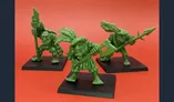 Three green ogre miniatures are displayed against a bright red background. Each figure is uniquely posed: one holds a large axe, another wields a spear, and the third carries a staff adorned with feathers. They stand on black bases, showcasing their detailed sculpting that pays homage to classic oldhammer aesthetics. The miniatures exhibit exaggerated features and playful expressions, embodying a whimsical yet menacing presence suitable for wargames and RPGs.