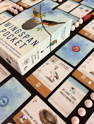A box labeled "Wingspan Pocket" with illustrations of birds and a nest on a branch. Surrounding the box are game cards featuring bird illustrations, eggs, and buttons. Some cards have visible text such as "AT LEAST 1 CARD TUCKED BEHIND IT", "STARTING HERE, ACTIVATE", and "PLAY A BIRD".