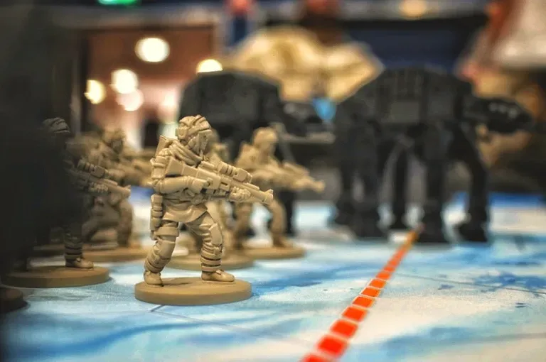 A group of miniature soldiers in beige uniforms, armed and poised for action, are positioned in the foreground on a tabletop battlefield. In the background, two large, black AT-AT walkers loom, creating a stark contrast against the lighter colors of the soldiers. The scene is set on a blue terrain mat, with a red line marking a path across the battlefield.