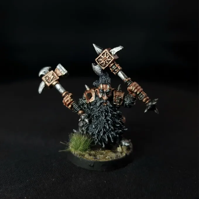 A miniature figure of a bearded dwarf-like character with long, dark hair and a thick beard, wearing detailed armor and holding two large, ornate hammers with intricate designs. The figure stands on a circular base with small patches of grass and textured ground. The background is black.