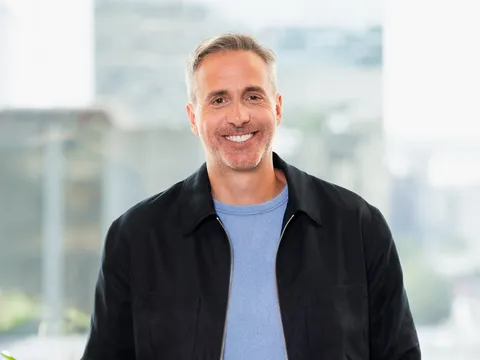 A man in a black jacket over a blue shirt, smiling at the camera. Office setting with modern glass windows and indoor plants in the background.
