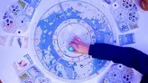 A hand points to a circular Arctic-themed board game map featuring constellations, wildlife icons, and labeled regions. Blue-toned illustration with symbols for animals, terrain, and game mechanics.