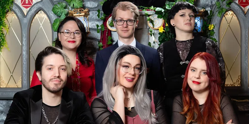 A group of six individuals poses together in front of a stone wall adorned with greenery. The front row features three people: a man with shoulder-length blonde hair and a black jacket, a woman with long, silver hair and glasses wearing a pink top, and another woman with dark hair styled in a bob, dressed in a black outfit. In the back row, a man with glasses stands between two women, one with dark hair and a red top, and the other with short black hair and a black dress. The overall atmosphere is vibrant and engaging, reflecting a sense of camaraderie.