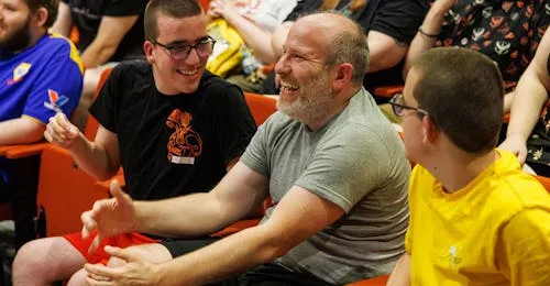 Three individuals are seated in a vibrant auditorium, engaged in a lively conversation. One person, wearing a black shirt with a graphic design, is laughing and gesturing animatedly, while another, in a gray shirt, smiles broadly, showing a friendly demeanor. The third individual, dressed in a yellow shirt, appears amused, contributing to the cheerful atmosphere of the gathering. The background features a diverse audience, indicating a lively event.