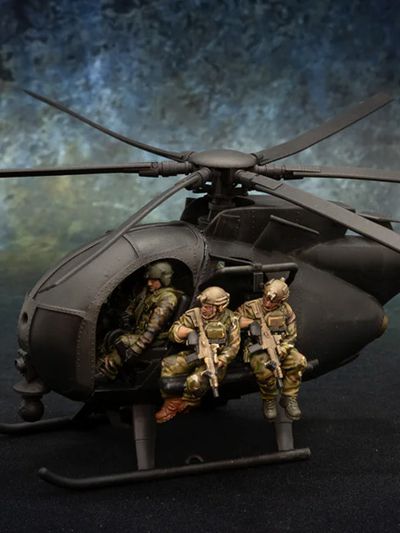 A detailed model of a Little Bird helicopter, featuring a dark color scheme, is prominently displayed. Two miniature soldiers are seated on the helicopter's skids, equipped with rifles, while another soldier is visible inside the cockpit. The background features a textured gradient, enhancing the focus on the helicopter and its crew.