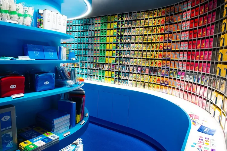 A vibrant display of Oink Games products fills a curved wall, showcasing a variety of colorful game boxes arranged in a grid pattern. The boxes feature bold designs in bright colors, including yellows, greens, blues, and pinks, creating an eye-catching visual effect. In the foreground, a blue shelf holds various game-related items and stationery, complementing the overall aesthetic of the store.