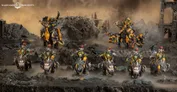 Miniature figures of six armored warriors with helmets and red plumes, holding weapons, standing on rocky terrain. One large armored figure on a mechanical beast raises a weapon. Background shows a ruined fortress with smoke and debris.