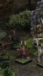 A group of intricately detailed fantasy miniatures stands in a lush, green setting near a stone ruin. The figures include various characters such as a skeletal leader holding a staff, a warrior with a bow, and a creature resembling a humanoid animal, all showcasing vibrant colors and classic 80s-style designs. Surrounding them are patches of grass and small bushes, enhancing the scene's immersive tabletop atmosphere.