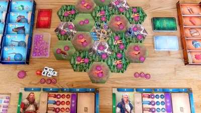 Board game setup with hexagonal green terrain tiles featuring pink and purple structures. Miniature figures and small tokens scattered across. Two player boards with character portraits, resource dials, and action icons. Dice and small bags of colored tokens on the table.