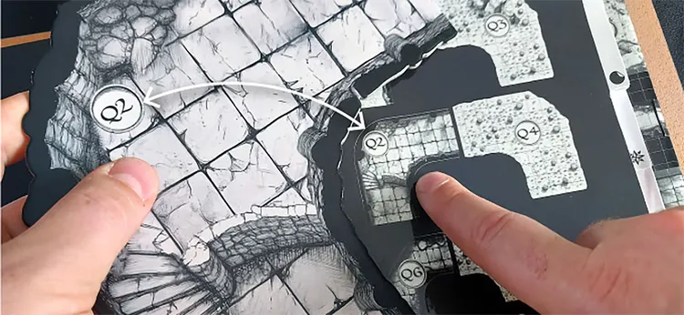 A close-up view of a modular mapping system for tabletop gaming, featuring a detailed, grimdark-style map with various sections labeled as Q2, R2, R4, R5, and R6. Two fingers are positioned on the map, indicating specific areas, while an arrow points from Q2 to R2, suggesting a connection or pathway. The textured surface of the map includes intricate stone patterns, enhancing the fantasy theme.