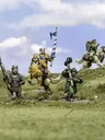 A group of halfling miniatures stands on a grassy terrain, showcasing their unique characteristics. In the center, a halfling in ornate armor rides a large, lion-like creature, holding a blue and white banner. To the left, another halfling wields a spear, while a third, dressed in green, brandishes a hammer. A fourth halfling rides a smaller, dog-like creature, completing the ensemble of pint-sized warriors. The background features a soft blue sky with scattered clouds, enhancing the whimsical atmosphere.