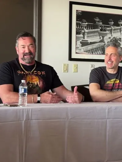 Two men are seated at a table during a panel discussion. On the left, a man with a beard, wearing a black shirt featuring a colorful graphic and a necklace, gestures with his hands while speaking. On the right, a man with short gray hair, dressed in a gray t-shirt with a retro design, smiles and listens attentively. Behind them, a framed black-and-white photograph of a decorative railing is visible on the wall. A bottle of water and a small device are placed on the table in front of them.