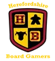 A shield-shaped logo features a divided design with four quadrants. The top left quadrant displays a red die with black dots, the top right features a black meeple, the bottom left shows a black cow silhouette, and the bottom right has a red die with white dots. The text "Herefordshire Board Gamers" is prominently displayed above and below the shield in a bold, yellow font.