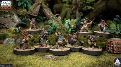 A group of four Ewok miniatures from the Star Wars: Legion game is displayed on circular bases, set against a lush, green forest backdrop. Each Ewok is uniquely posed: one holds a spear, another brandishes a slingshot, while the others are equipped with various weapons and accessories. The terrain features rocks, foliage, and remnants of a crashed vehicle, enhancing the scene's immersive quality.