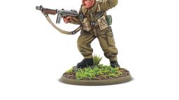 A detailed miniature of a soldier in a military uniform, standing on a circular base adorned with grass and dirt. The figure, representing Augustus Newman VC, is depicted in a dynamic pose, holding a submachine gun in one hand while raising the other in a commanding gesture. He wears a green beret and a khaki uniform, complete with various pouches and gear attached to his chest.