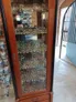 A wooden display cabinet filled with a vast array of painted miniatures, showcasing various fantasy figures and models. The cabinet has multiple shelves, each densely packed with detailed miniatures, including warriors, creatures, and vehicles, reflecting a rich history of tabletop gaming. In the background, a glimpse of the shop's interior and an open door can be seen, adding to the atmosphere of the Foundry's headquarters.