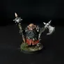 A painted miniature figure of a bearded dwarf-like warrior with dark skin, wearing a red and black outfit with metallic armor details. The figure holds a large double-headed axe with spiked ends in one hand and a spiked mace in the other. The base has small patches of green grass-like texture. The background is black.