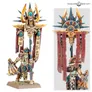 Miniature of a fantasy warrior kneeling, holding a large ornate staff topped with a sunburst design and a skull emblem. Wears elaborate armor with gold, blue, and red details, including a headdress with a blue gem. Standing on a rectangular base with grass texture.