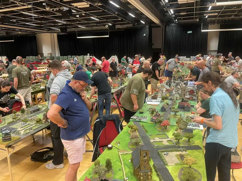A bustling scene at the Britcon Historical Wargaming Convention, held in a spacious venue filled with tables showcasing intricate wargaming setups. Attendees, both men and women, are engaged in gameplay and discussions, surrounded by detailed miniature landscapes, buildings, and figures. The atmosphere is lively, with a mix of focused players and social interactions, highlighting the community aspect of the event.