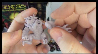 A person holds a detailed miniature of a creature from Fenris Games' Wyrdworld series, showcasing its intricate features such as muscular limbs and a fierce expression. The miniature is unpainted and made of a light-colored material, emphasizing its sculpted details. In the background, a colorful game box titled "GENESIS: The First Book of Nightmares" is partially visible, along with other miniatures displayed on a shelf.