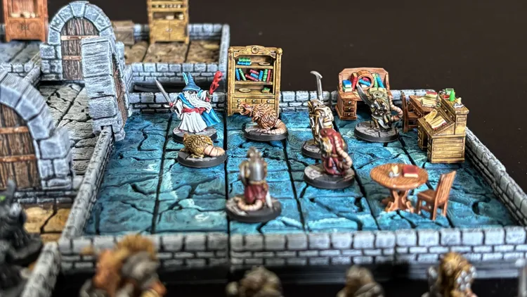 A detailed 15mm scale dungeon scene features a blue-tiled floor with various miniatures, including a wizard in a blue robe wielding a sword, and several ratmen. The dungeon is adorned with furniture pieces like a bookshelf filled with books and a workbench, set against stone walls. The modular design includes multiple tiles and scattered items, creating an immersive tabletop environment.