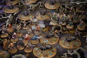 A detailed tabletop scene features various miniatures from the Middle-earth Strategy Battle Game, depicting a tense moment in Mirkwood. Prominent are several large spiders on circular bases, surrounded by a mix of characters including Bilbo, Thorin, and other dwarves, all positioned on a textured terrain with rocks and twisted roots. The dark, earthy colors of the setting enhance the atmosphere of the iconic "Flies and Spiders" chapter from The Hobbit.