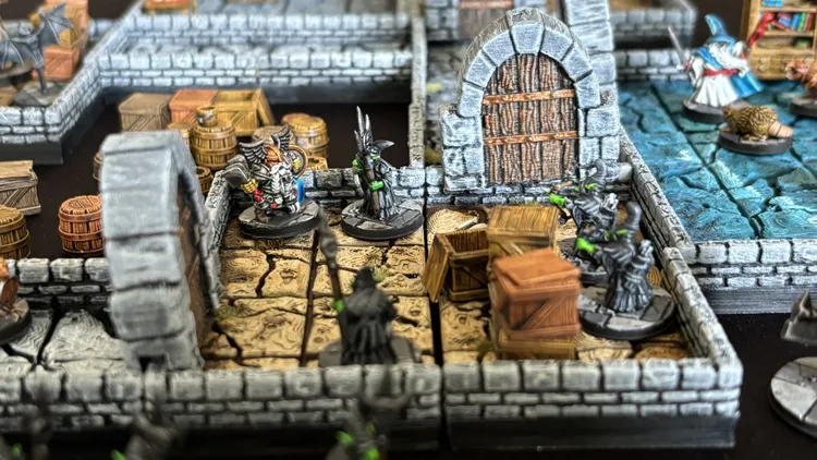 A detailed tabletop scene features a modular dungeon made up of stone walls and cracked floor tiles. In the foreground, two miniatures stand: a heavily armored hero with a shield and a staff-wielding figure in dark robes. Surrounding them are wooden crates and barrels, enhancing the dungeon's immersive atmosphere.