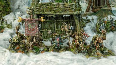 A group of intricately detailed miniatures inspired by Celtic mythology is displayed in front of a rustic, weathered structure covered in moss and foliage. The miniatures, depicting various fantastical creatures, include a central figure holding a banner, flanked by others wielding weapons and adorned with unique features like tusks and intricate patterns. The scene is set amidst a misty, ethereal atmosphere, enhancing the mystical theme of the Famhairean campaign.