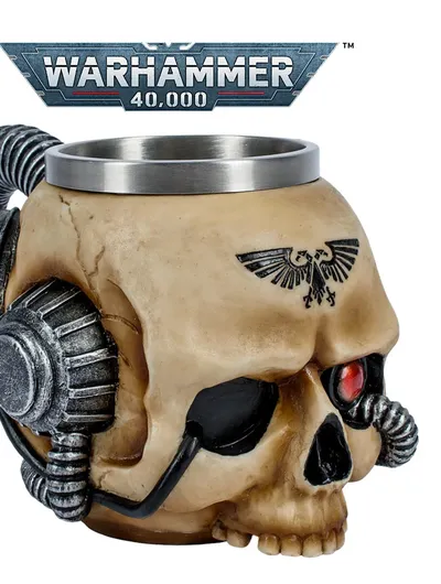 Beige ceramic mug shaped like a stylized human skull with mechanical augmentations. A metal rim lines the top of the mug. Two large, cylindrical, metallic mechanical components resembling pistons or servos are attached to the sides of the skull, connected by coiled black cables. A black eagle emblem with outstretched wings and a skull in the center is tattooed or printed on the forehead. The background shows a metallic banner with the text "WARHAMMER 40,000" in bold, futuristic lettering. The banner has a red and white stripe along the bottom edge.