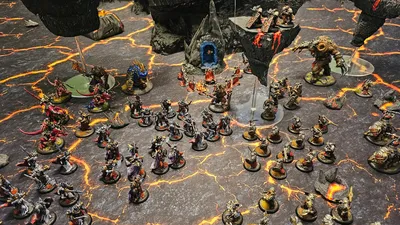 Miniature dark fantasy wargame scene with two armies on a cracked, lava-like battlefield. Left side features dwarven figures with armor and weapons, including a large blue-robed figure with a glowing orb.