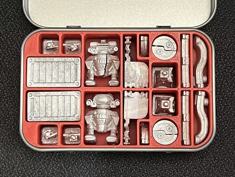 A small, silver tin is open to reveal a neatly organized assortment of miniatures and terrain pieces for the Space Denizens wargame. The contents include two distinct mechs, various infantry figures, and several terrain features such as crates and circular components, all set against a bright red insert. The miniatures are metallic and intricately detailed, showcasing the charm of the 8mm scale.