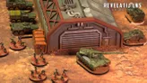 A detailed tabletop scene features a large, industrial building with a corrugated roof and a prominent garage door, set against a textured, sandy ground. In the foreground, several infantry miniatures are arranged in a semi-circle, while a green tank is positioned nearby, showcasing intricate details. Behind the building, additional vehicles are lined up, contributing to the military theme of the setting, with the "Revelations: Skirmish" logo visible in the top right corner.