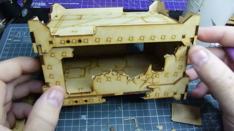 A partially assembled, light brown cardboard or MDF terrain piece with a damaged, jagged top edge, revealing an open interior. The structure has square and rectangular cutouts, with small square and circular holes along the edges. It rests on a green self-healing cutting mat with a grid pattern. A hand is visible on the left side, holding part of the terrain. Small debris and tools, including a metal L-shaped piece, are scattered on the mat.
