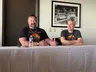 Two men are seated at a table during a panel discussion. On the left, a man with a beard, wearing a black shirt featuring a colorful graphic and a necklace, gestures with his hands while speaking. On the right, a man with short gray hair, dressed in a gray t-shirt with a retro design, smiles and listens attentively. Behind them, a framed black-and-white photograph of a decorative railing is visible on the wall. A bottle of water and a small device are placed on the table in front of them.