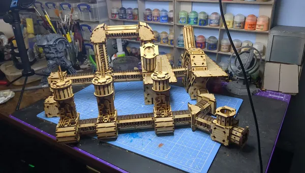 A detailed assembly of the TT Combat Sector 4 Plasma Relay is displayed, featuring intricate wooden structures with multiple towers and connecting pathways. The design includes cylindrical towers with circular tops and various platforms, all set against a blue cutting mat. In the background, jars of paint and tools are visible, indicating a workspace dedicated to tabletop gaming and model building.