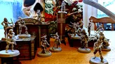 A collection of detailed miniatures from the game "Boarders and Black Flags" is displayed on a wooden surface, featuring both human pirates and undead Skelliwags. The miniatures are unpainted and vary in poses, with some wielding swords and others appearing to engage in action. In the background, a vibrant pirate-themed backdrop showcases a skull and crossbones design, along with elements of a ship, enhancing the thematic atmosphere of the game.