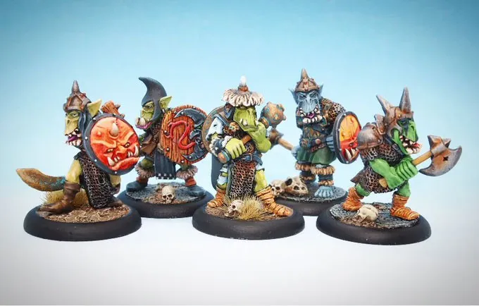 A group of five intricately painted orc miniatures stands on a textured black base, showcasing a variety of weapons and armor. Each orc features distinct details, such as shields with unique designs, including a fierce red face, and various outfits that highlight their individual characteristics. The background is a soft gradient of blue, enhancing the vibrant colors of the miniatures.