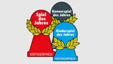 Three award trophies are displayed, each representing a different category of the Spiel des Jahres. The red trophy, labeled "Spiel des Jahres," features a laurel wreath design, symbolizing the Game of the Year. The gray trophy, labeled "Kennerspiel des Jahres," represents the Connoisseur Game of the Year, while the blue trophy, labeled "Kinderspiel des Jahres," signifies the Children's Game of the Year. All trophies are marked with the text "KRITIKERPREIS" at the base.
