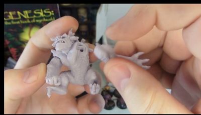A person holds a detailed miniature of a creature from Fenris Games' Wyrdworld series, showcasing its intricate features such as muscular limbs and a fierce expression. The miniature is unpainted and made of a light-colored material, emphasizing its sculpted details. In the background, a colorful game box titled "GENESIS: The First Book of Nightmares" is partially visible, along with other miniatures displayed on a shelf.