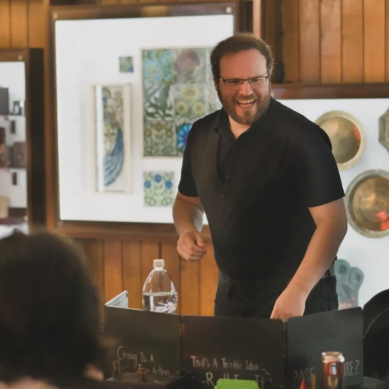 A smiling game master stands behind a black game screen, engaging with players at a tabletop gaming event. He wears a black shirt and glasses, with a water bottle on the table beside him. The background features wooden walls adorned with various artworks and decorative plates, creating a warm, inviting atmosphere for the gaming session.