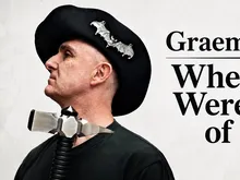 Man in black beret with bat pins, wearing a metal collar with coiled cord, black shirt.
