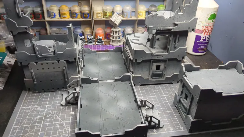 A tabletop model of a ruined multi-level industrial or sci-fi building made of gray plastic or resin pieces, with detailed walls, walkways, railings, and open doorways. On the table, there are three small painted miniature figures, a tall white plastic tower structure, and a purple grid mat partially visible beneath the model. In the background, shelves hold numerous small jars and pots of paint, with visible labels including "Citadel," "Layer," "Base," "Terminatus Stone," and "Lahmian Medium." The scene is set on a green cutting mat with measurement markings.