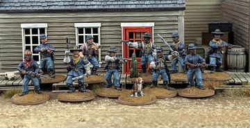 A group of ten detailed 28mm scale miniatures representing dismounted US Cavalry and Buffalo Soldiers stands in front of a wooden building with a red door. The miniatures are posed with various weapons, including rifles and a bugle, showcasing a mix of uniforms and accessories, such as scarves and hats. A small beagle and a cactus are also present among the figures, all mounted on 25mm round bases with a sandy terrain effect.