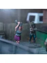 A dynamic scene features three miniature figures in a tense urban setting. In the foreground, a woman with blue hair raises a shovel, while a second figure behind her holds a baseball bat, both appearing ready to confront a threat. A third figure, dressed in a light shirt and blue pants, is seen running away, adding to the sense of urgency in the scene, with a green dumpster and a wooden fence in the background.