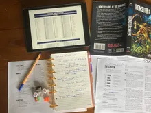 Topdown photo of a solo game in progress. Roll table from Mythic on tablet, Monster of the Week rulebook, journal for note taking, reference sheets