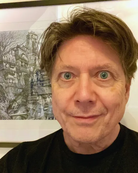 A close-up portrait of a man with light brown hair and striking green eyes, smiling slightly. He is wearing a black shirt and is positioned in front of a detailed, artistic background featuring a complex, fantastical structure, possibly a building or landscape. The overall tone of the image is casual and inviting, with a focus on the subject's expressive features.